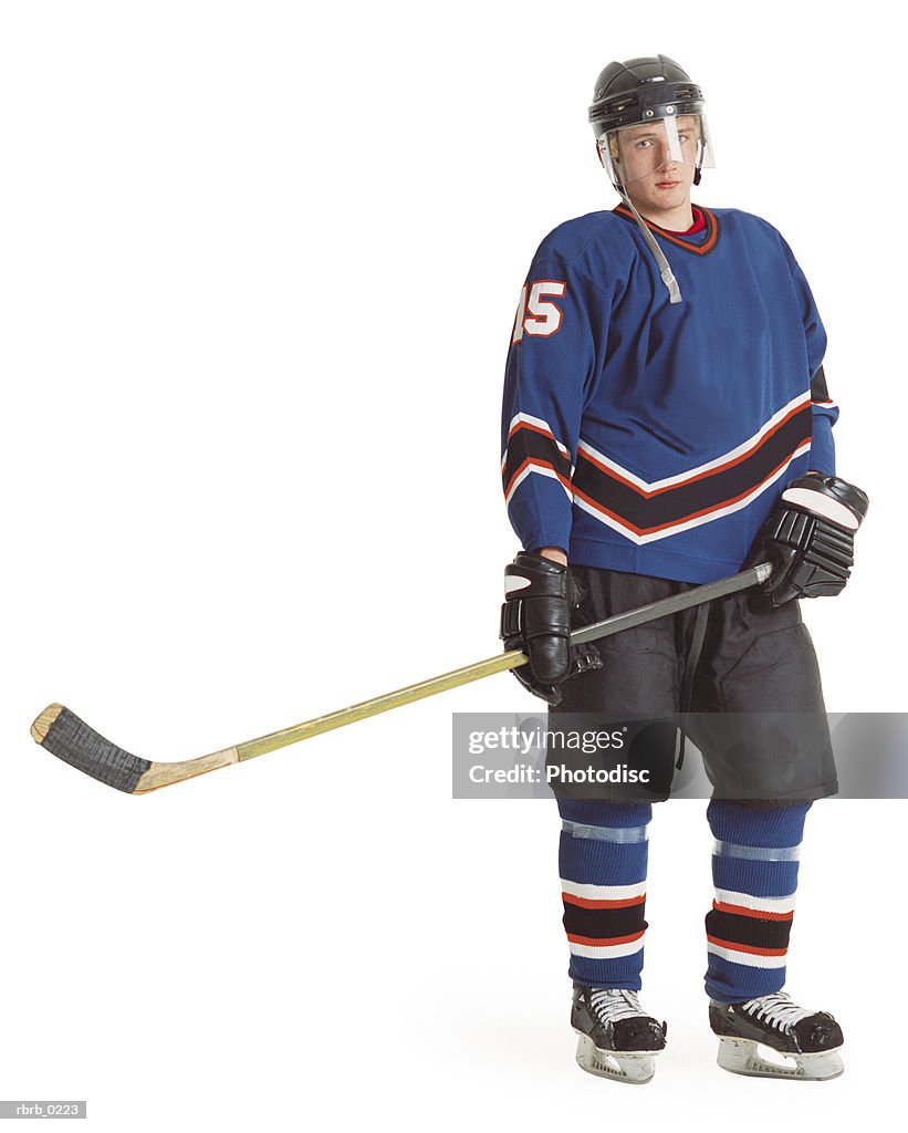 An adult caucasian male hockey player in a blue jersey stands with his stick extended and displays a stern look