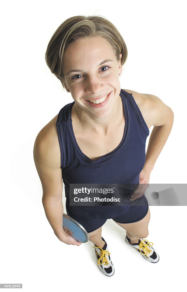 A young caucasian female track and field participant in a blue tank and shorts stands with a discus looking up at the camera