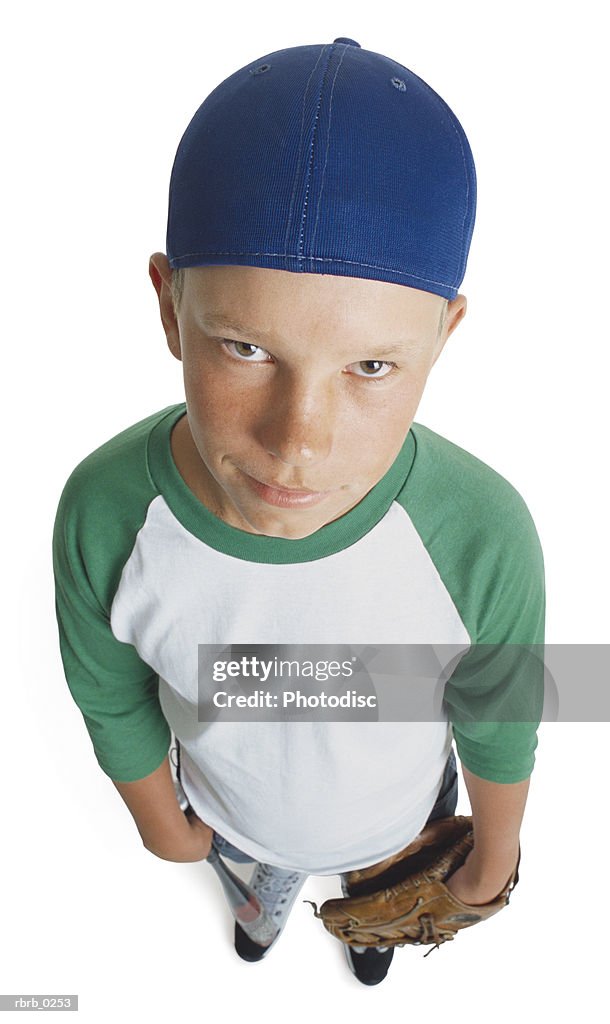 A caucasian male preteen in a green and white shirt and blue cap holds his mitt and smirks looking up towards the camera