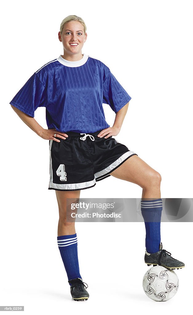 A young blond caucasian female soccer player in a blue jersey and black shorts poses smiling with her foot on the soccer ball