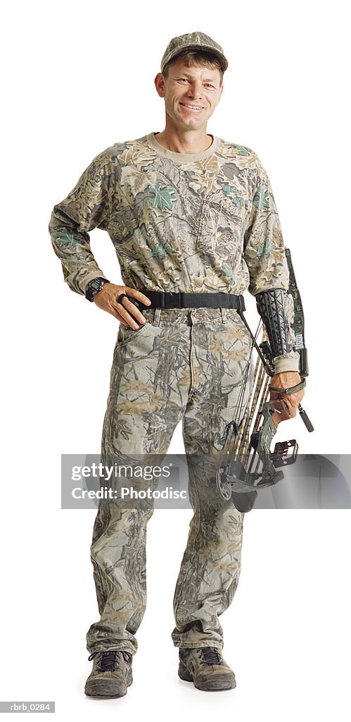 A caucasian man wearing army fatigues is standing and holding his archery equipment with his hand on his hip