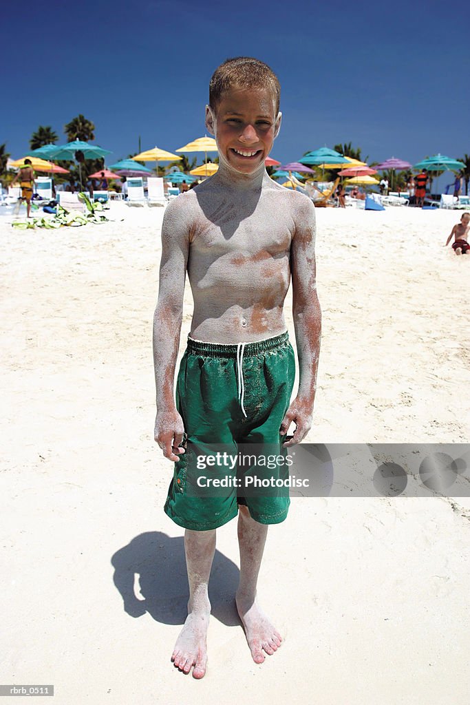A caucasian male child in a green swimsuit smiles as he plays in sand at a beach