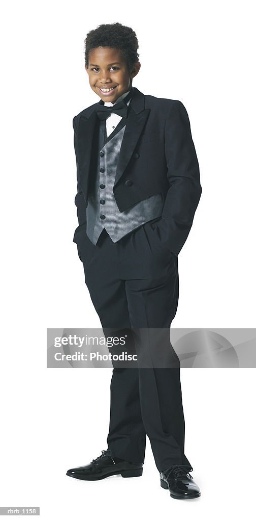 An african american male child in a tuxedo puts his hands in his pockets and smiles