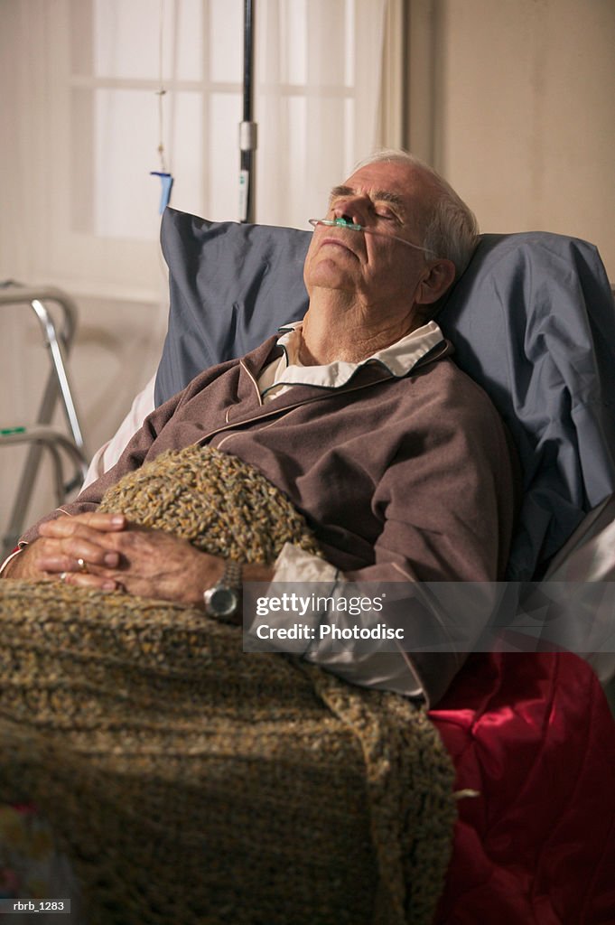 An elderly caucasian man in a brown robe lays in bed and rests with the aid of an oxygen tube