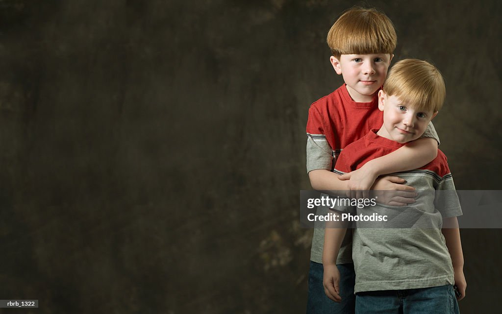 Studio portrait of a caucasian redheaded male child as he puts his arms around his little brother as they smile