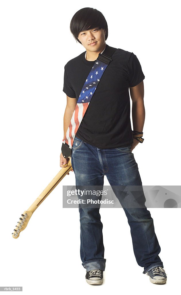 An asian teenage male in jeans and a black shirt smiles as he holds his electric guitar