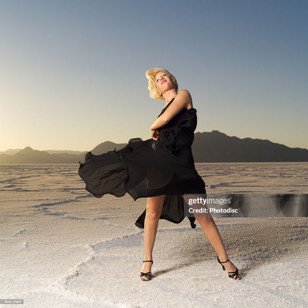 A young blonde woman dressed in a black dress tosses her skirt in a desert setting