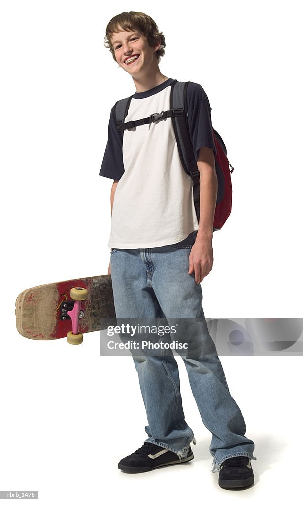 A caucasian male teen in jeans and a white and blue shirt stands with his backpack and skateboard