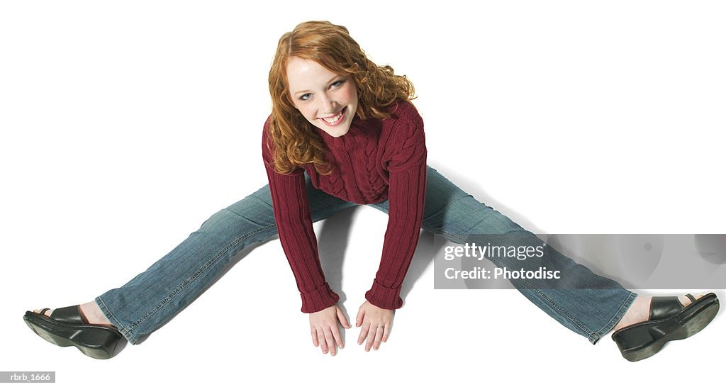 A caucasian redheaded female teen in jeans and a red sweater spreads her legs out and smiles up at the camera
