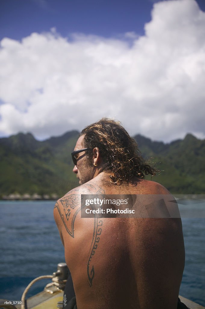 A caucasian man with a tattoo rides on a boat while vacationing in the tropics