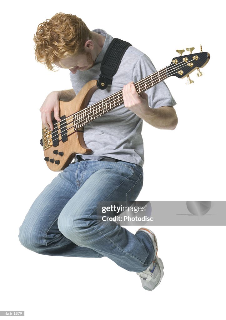 Full body shot of a young adult male in a grey shirt as he jumps up with his guitar