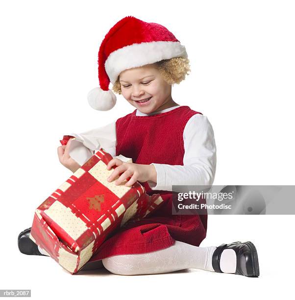 full body shot of a female child in a red dress and santa hat as she unwraps a present - torn gift stock pictures, royalty-free photos & images