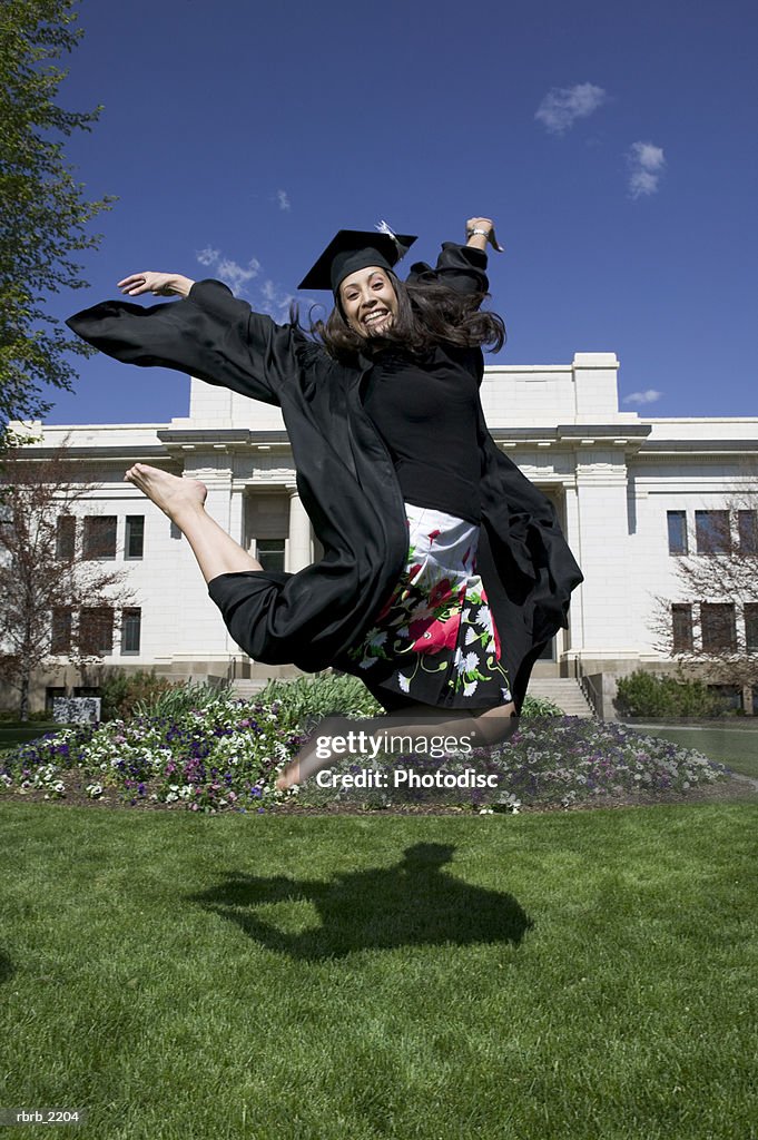 Full length wide shot of a young adult female college graduate as she excitedly jumps through the air