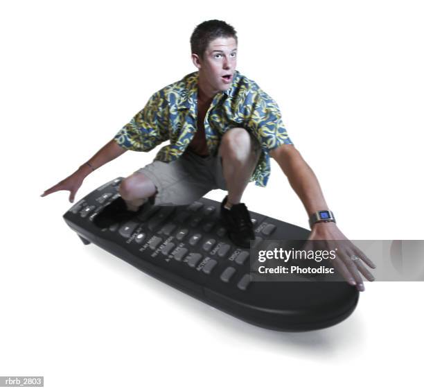 conceptual shot of a teenage male in a hawaiian shirt as he surfs on a tv remote control - control photos et images de collection