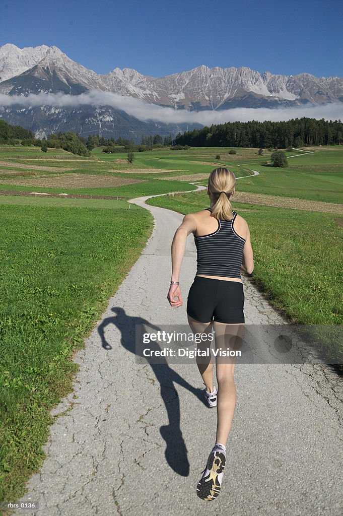 Wide shot of a young female jogger as she runs down a long path near a mountain range