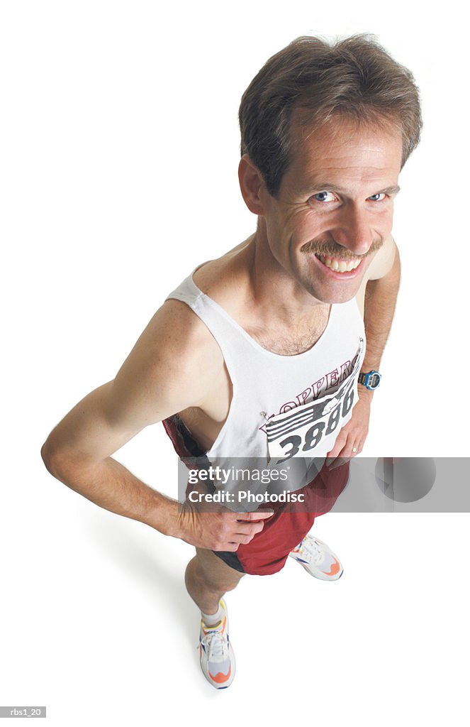 A caucasian man with a mustache is wearing red running shorts and a marathon number as he stands looking up at the camera