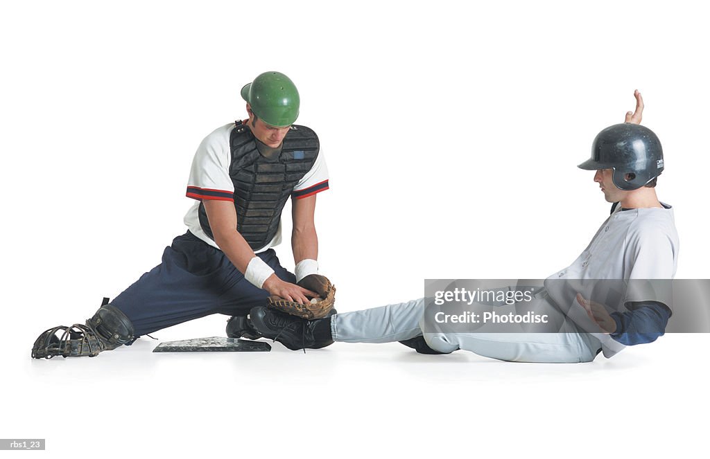 Two caucasian men are dressed in baseball uniforms as one in a catcher vest tags the other wearing a batter helmet out