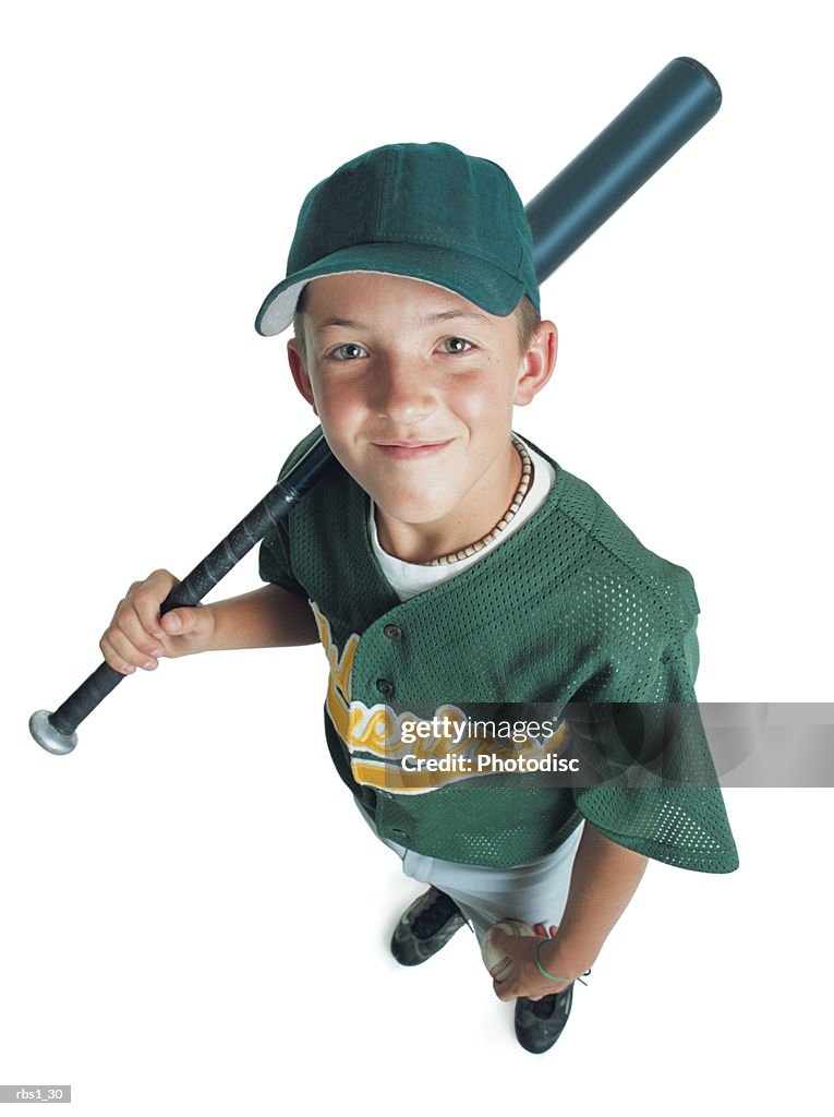 A young caucasian boy is wearing a green little league uniform and holding a bat over his shoulder as he smiles up at the camera