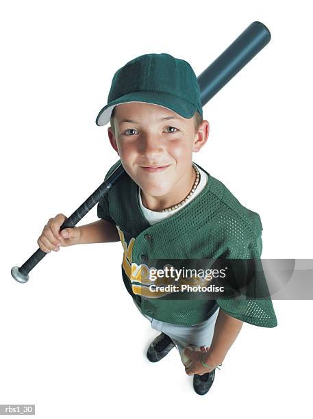 a young caucasian boy is wearing a green little league uniform and holding a bat over his shoulder as he smiles up at the camera - sports uniform stock pictures, royalty-free photos & images