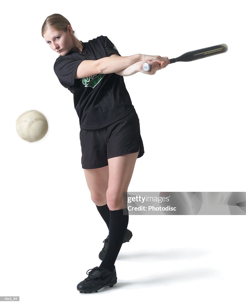 A pretty young caucasian woman is wearing a black softball uniform and swinging her bat after hitting a ball
