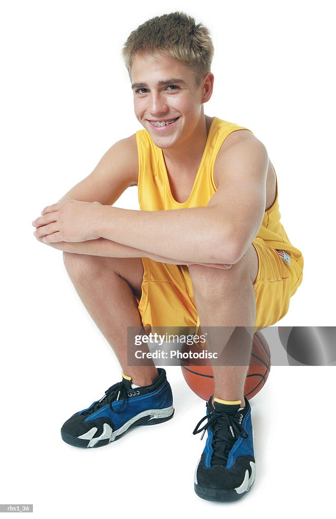 A caucasian teenage boy is wearing a yellow basketball uniform and smiling as he sits on his basketball
