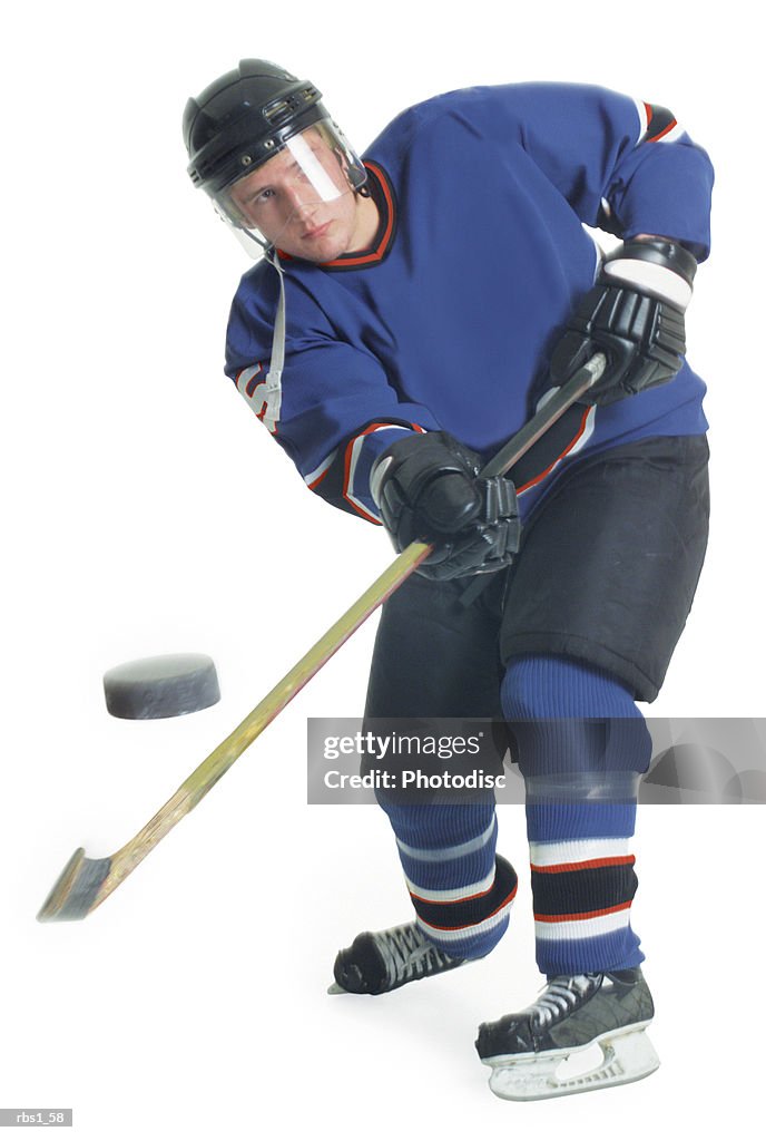 A young caucasian male hockey player in a blue uniform shoots the puck towards the camera with his hockey stick