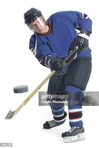 a young caucasian male hockey player in a blue uniform shoots the puck towards the camera with his hockey stick - ice hockey uniform stock pictures, royalty-free photos & images