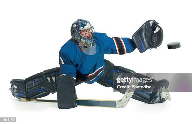 a caucasian male hockey goalie in a blue uniform splits his legs and reaches for the puck to block a shot - difensore hockey su ghiaccio foto e immagini stock