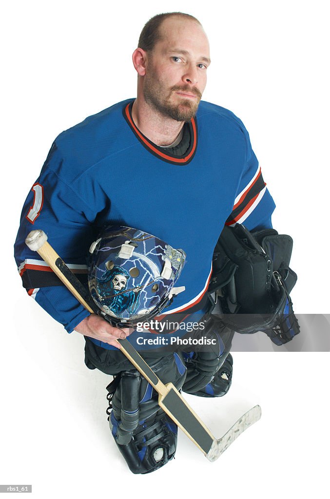 An adult bearded caucasian male hockey player holds his equipment and smiles smugly as he looks up at the camera
