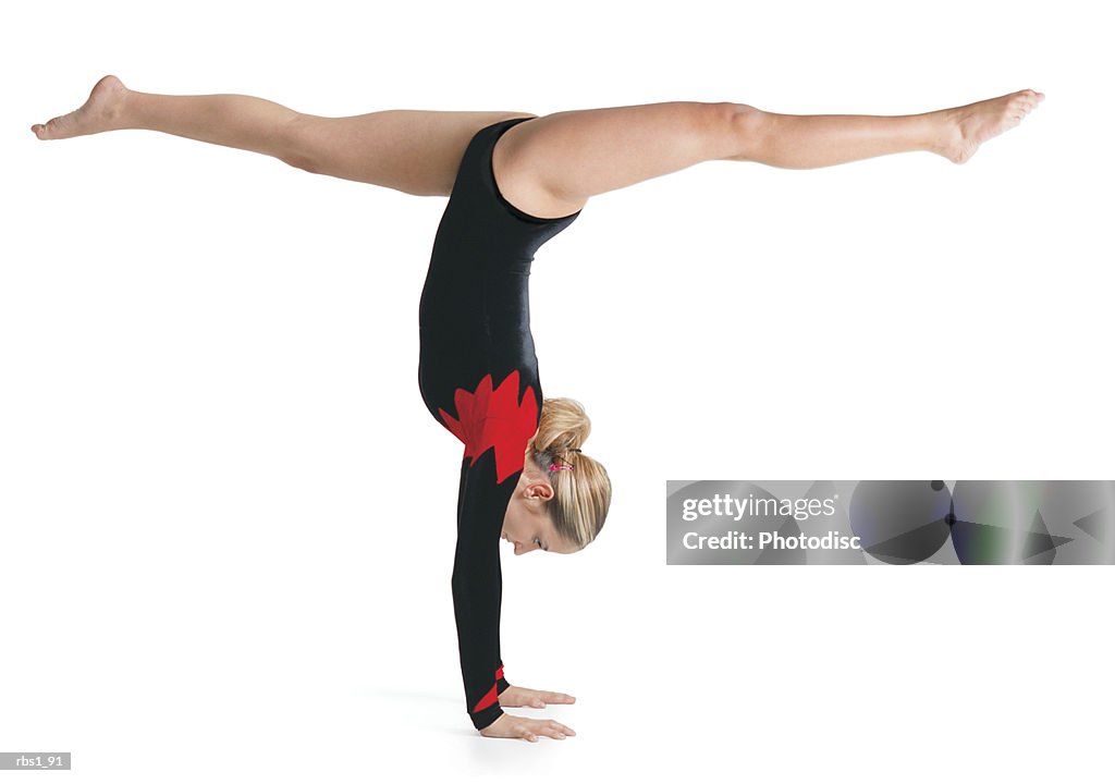 A young caucasian female gymnist in a red and black leotard does a handstand while doing the splits with her legs