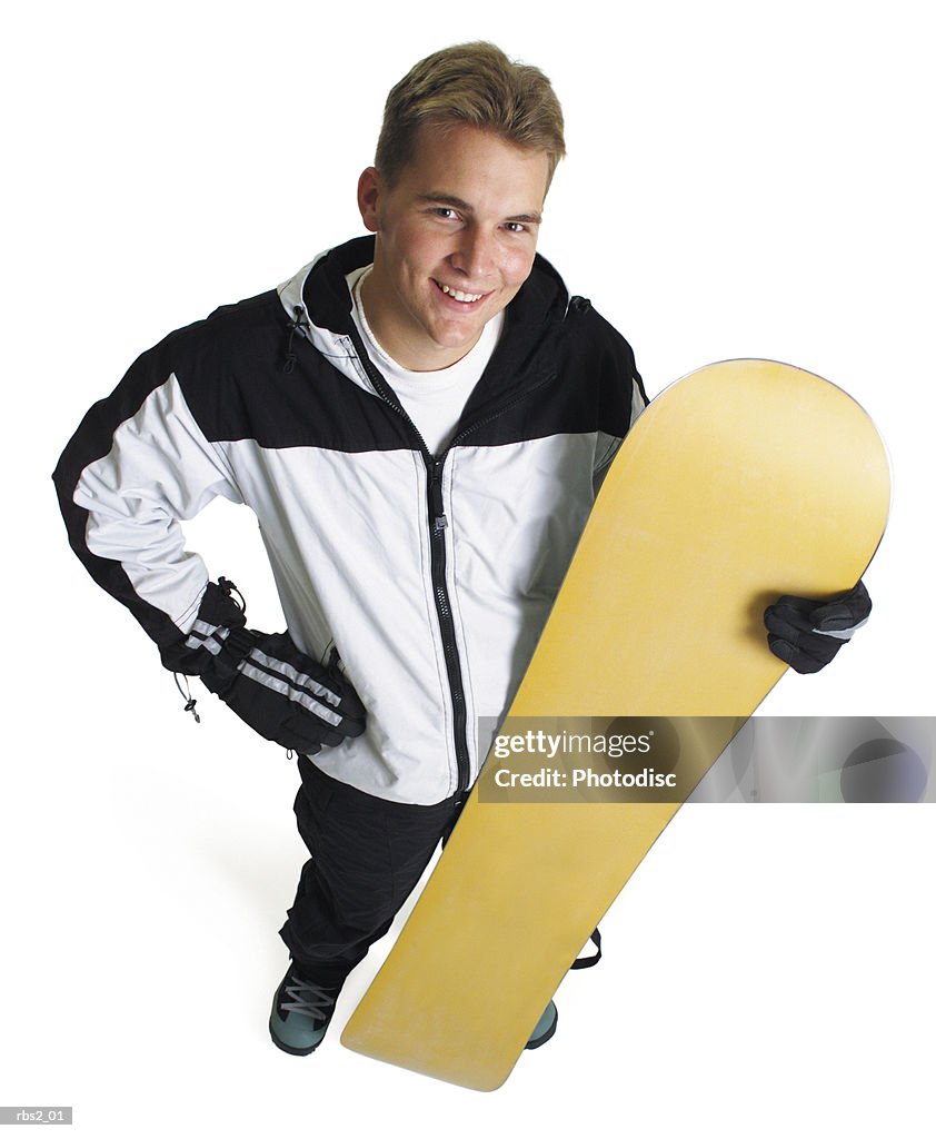 A young caucasian male snowboarder stands holding his board and smiles as he looks up at the camera
