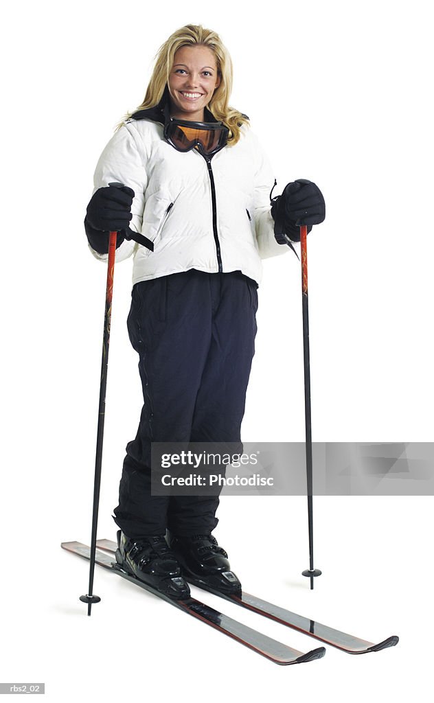 A young caucasian female skier in a white coat stands smiling with her snow skis and poles