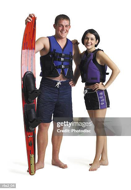 a young caucasian couple stand in lifejackets and swimming suits as the male holds a waterski and the two friends smile - equipo de seguridad para el mar fotografías e imágenes de stock