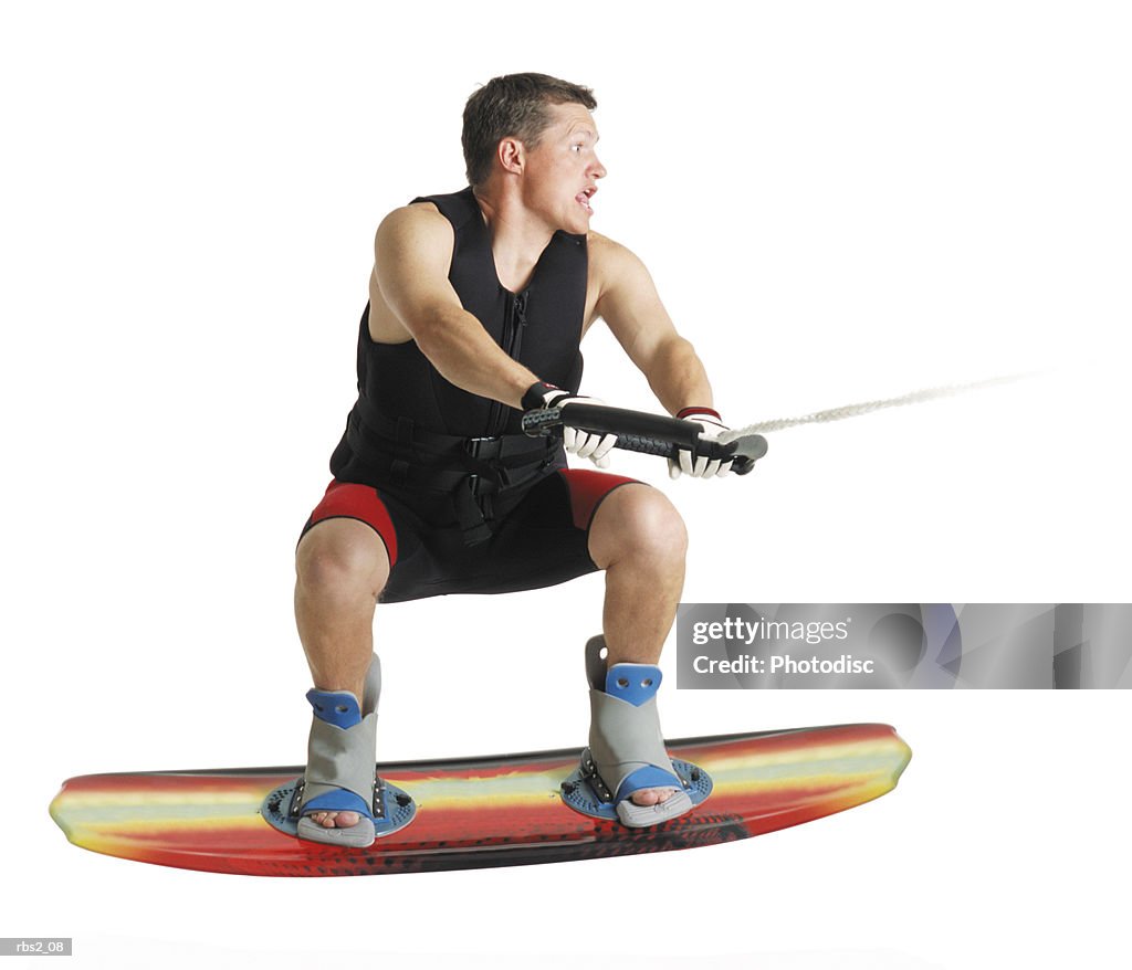 A caucasian male wakeboarder in a black lifejacket stands gripping the tow line and jumping in the air on his board