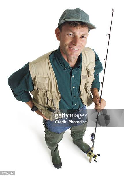 an adult caucasian male fisherman in a green shirt and tan fishing vest stands holding his pole and smiling as he looks up into the camera - rod stock pictures, royalty-free photos & images