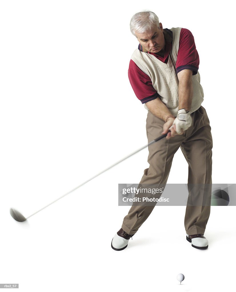 A middle age man with white hair is wearing a tan vest over a red golf shirt as he swings at a golf ball on a tee