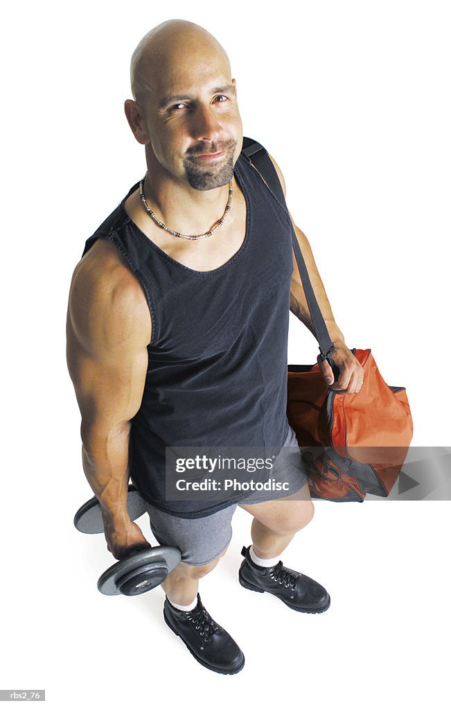 An adult caucasian male weightlifter in a black tank top holding a dumbell and his gear stands and looks up at the camera