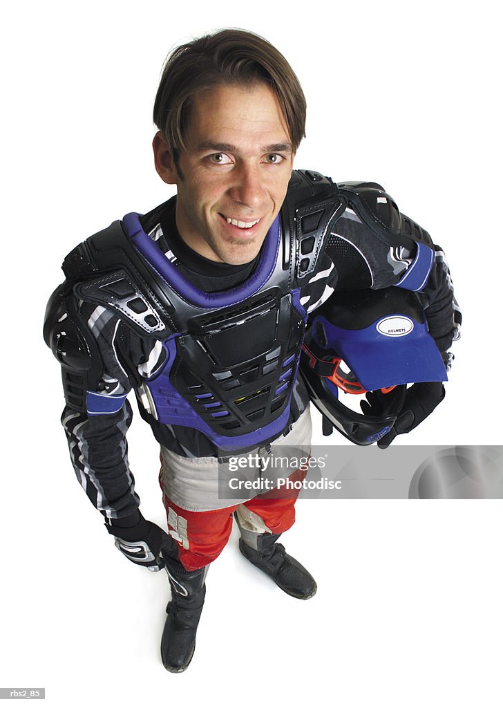A young caucasian male dirtbiker holds his helmet under his arm and smiles up into the camera