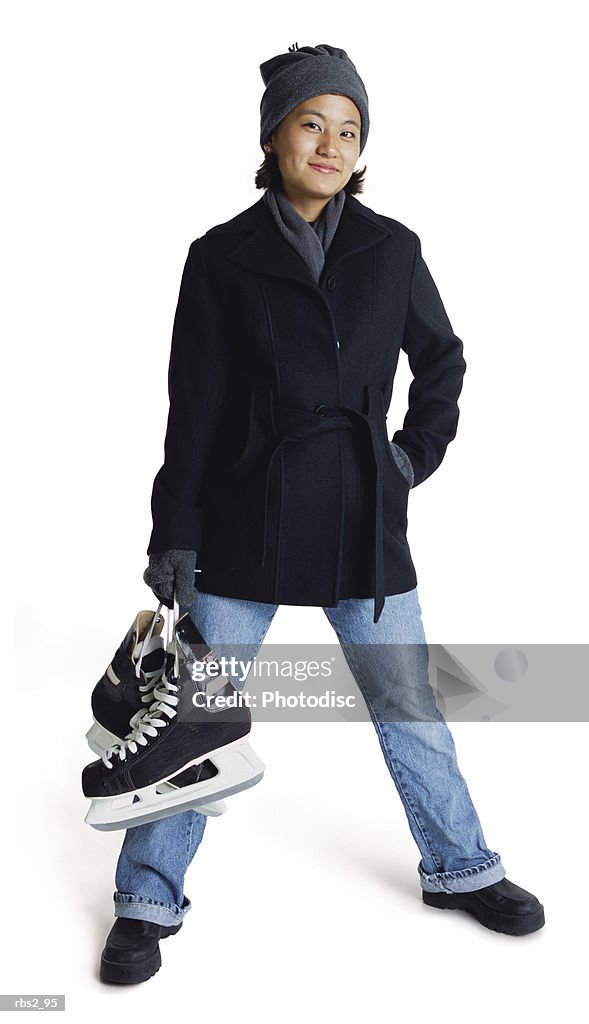 An asian teenage girl wearing a winter coat and hat is holding her ice skates and standing spread legged