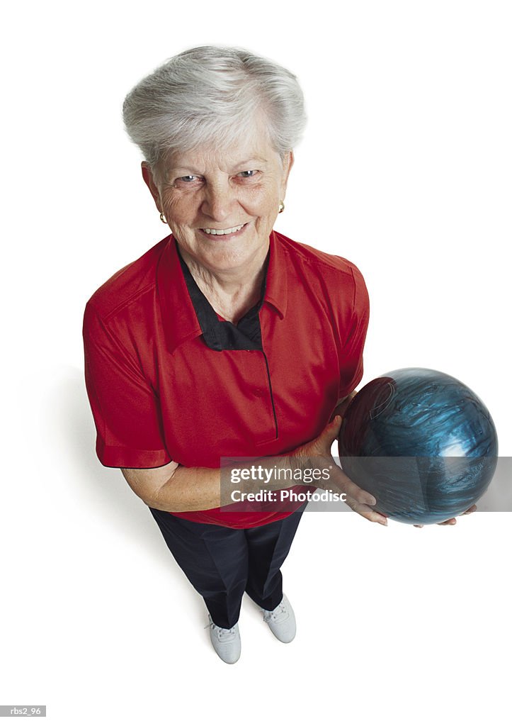 An elderly caucasian woman with white hair is wearing a red bowling shirt and holding her blue bowling ball as she looks up at the camera smiling