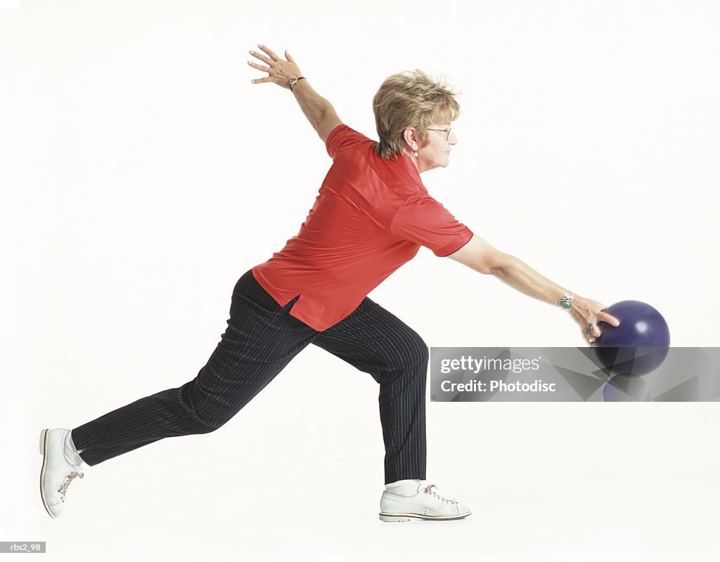 An elderly caucsian woman with light brown hair wearing a red bowling shirt is running forward about to throw her blue bowling ball