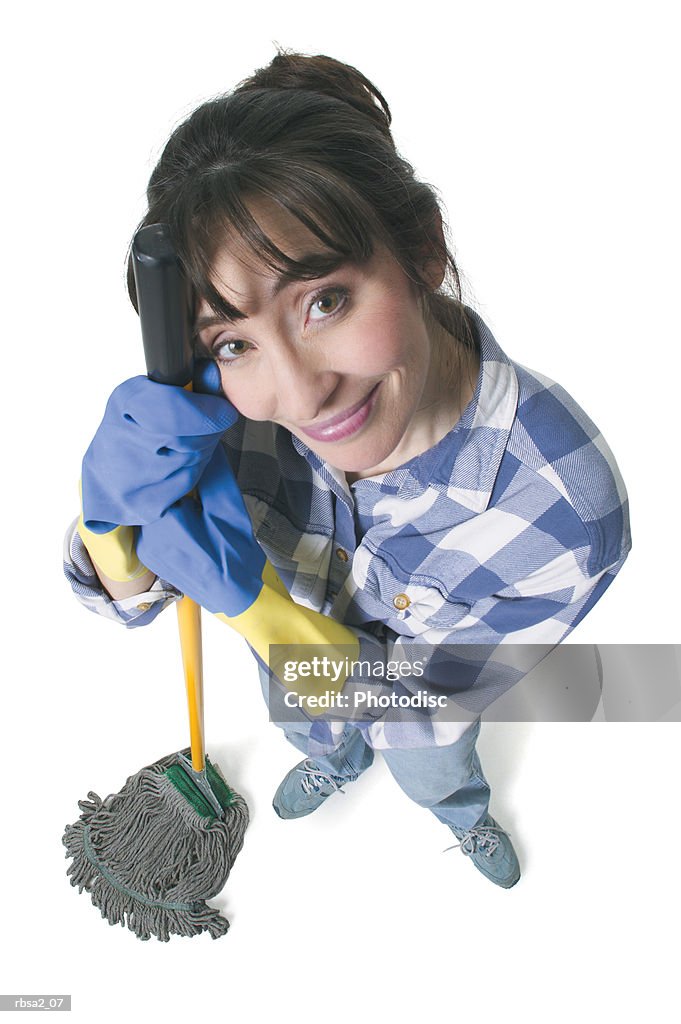 A caucasian woman in jeans and a plaid shirt wears rubber gloves and holds a mop as she looks up into the camera