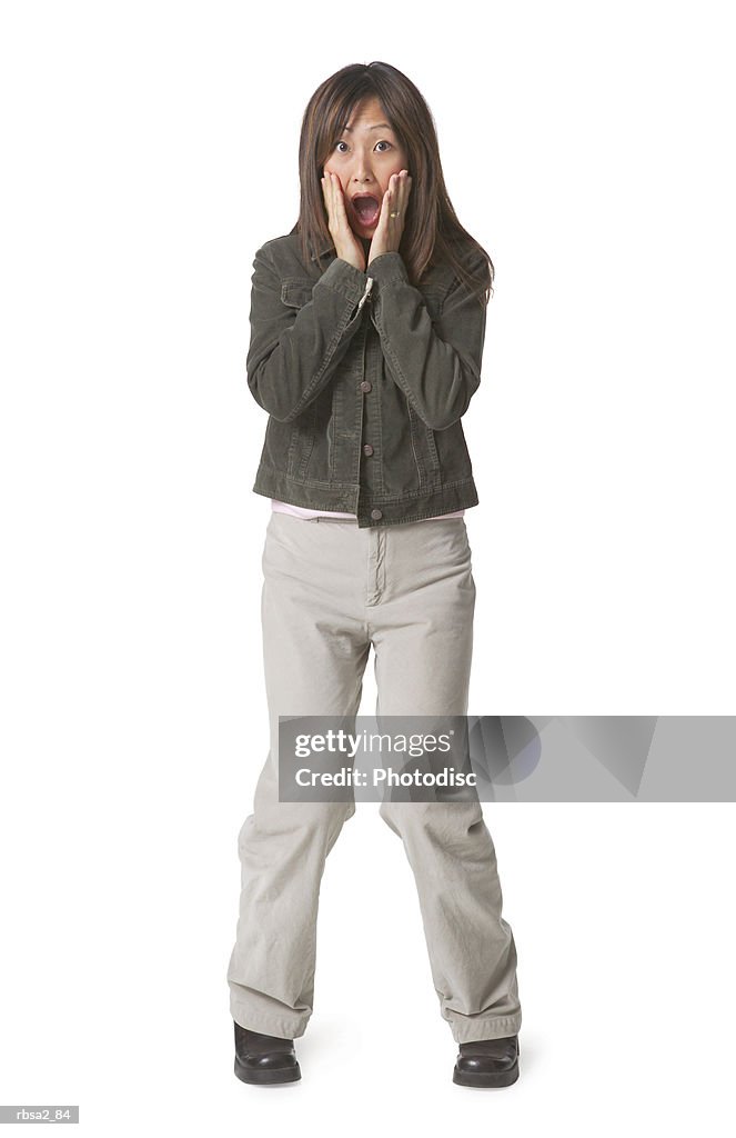 A young asian woman dressed in tan pants and a jean jacket puts her hands on her face and acts shocked and surprised