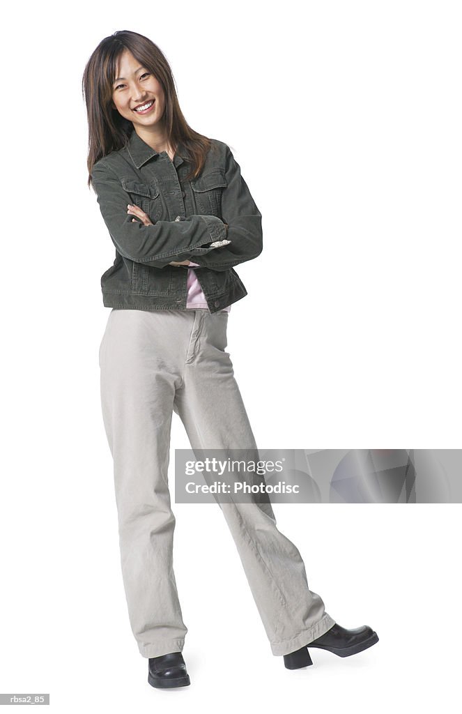 A young asian woman dressed in tan pants and a jean jacket folds her arms leans to one side and smiles