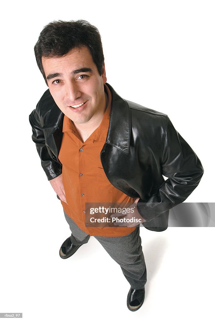 An adult caucasian man in an orange shirt and leather jacket puts his hands on his hips and smiles up to the camera