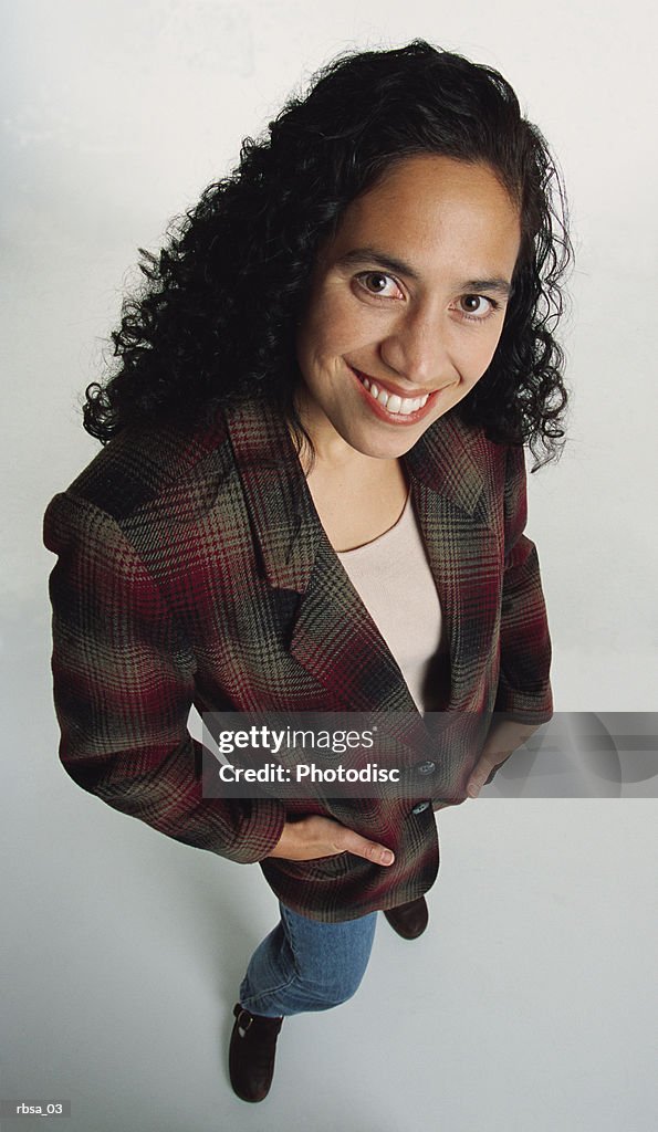 Beautiful young ethnic adult female with long dark curly hair wearing a checked blazer stands and looks up at the camera while smiling approvingly