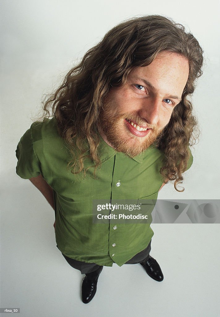 Young caucasian adult male hippie with facial hair and long curly hair wears a green shirt and stands looking up at the camera with a mischievous smile