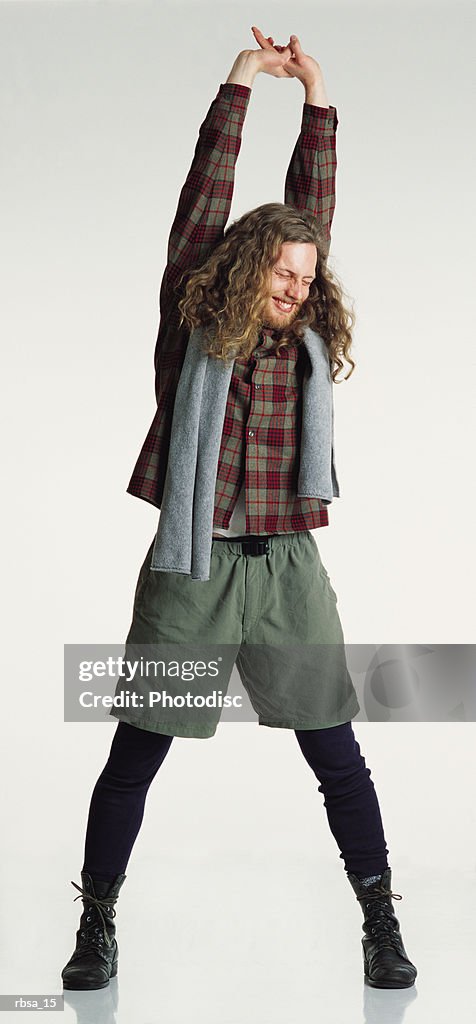 Smiling young caucasian adult male hippie with long curly hair wearing a checkered shirt and khaki shorts with a towel over his shoulders stands stretching his arms over his head