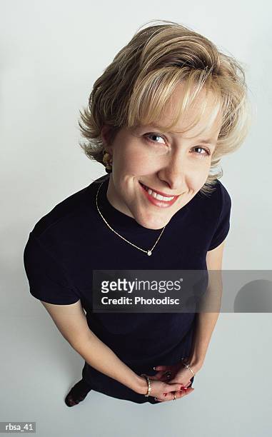 attractive young blonde caucasian adult female with short hair wearing a dark dress and diamond necklace stands looking up at the camera with a pretty smile - smile stockfoto's en -beelden