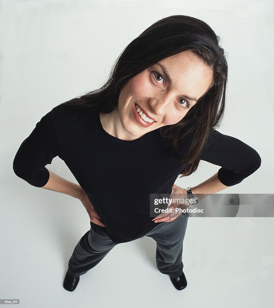 Beautiful young bruenette caucasian adult female wearing dark slacks and a dark shirt has her hands on her hips as she cocks her head and looks up at the camera smiling